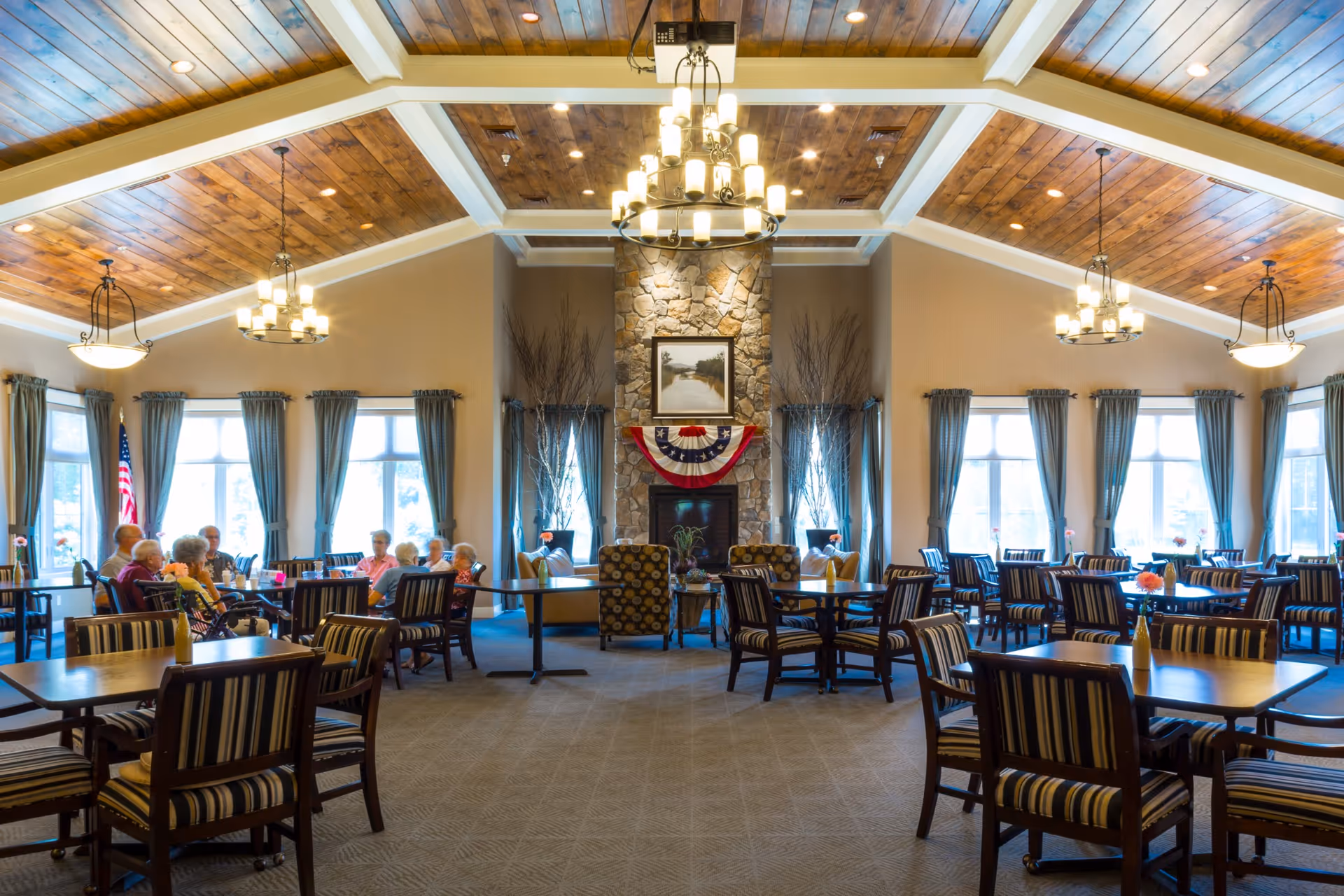 A spacious dining room with wooden tables and striped cushioned chairs arranged neatly. Several elderly people are seated at one of the tables near large windows with blue curtains. The room features a high wooden ceiling with exposed beams and multiple chandeliers. A stone fireplace decorated with a patriotic banner and a framed painting is centered on the far wall, flanked by tall vases with decorative branches.