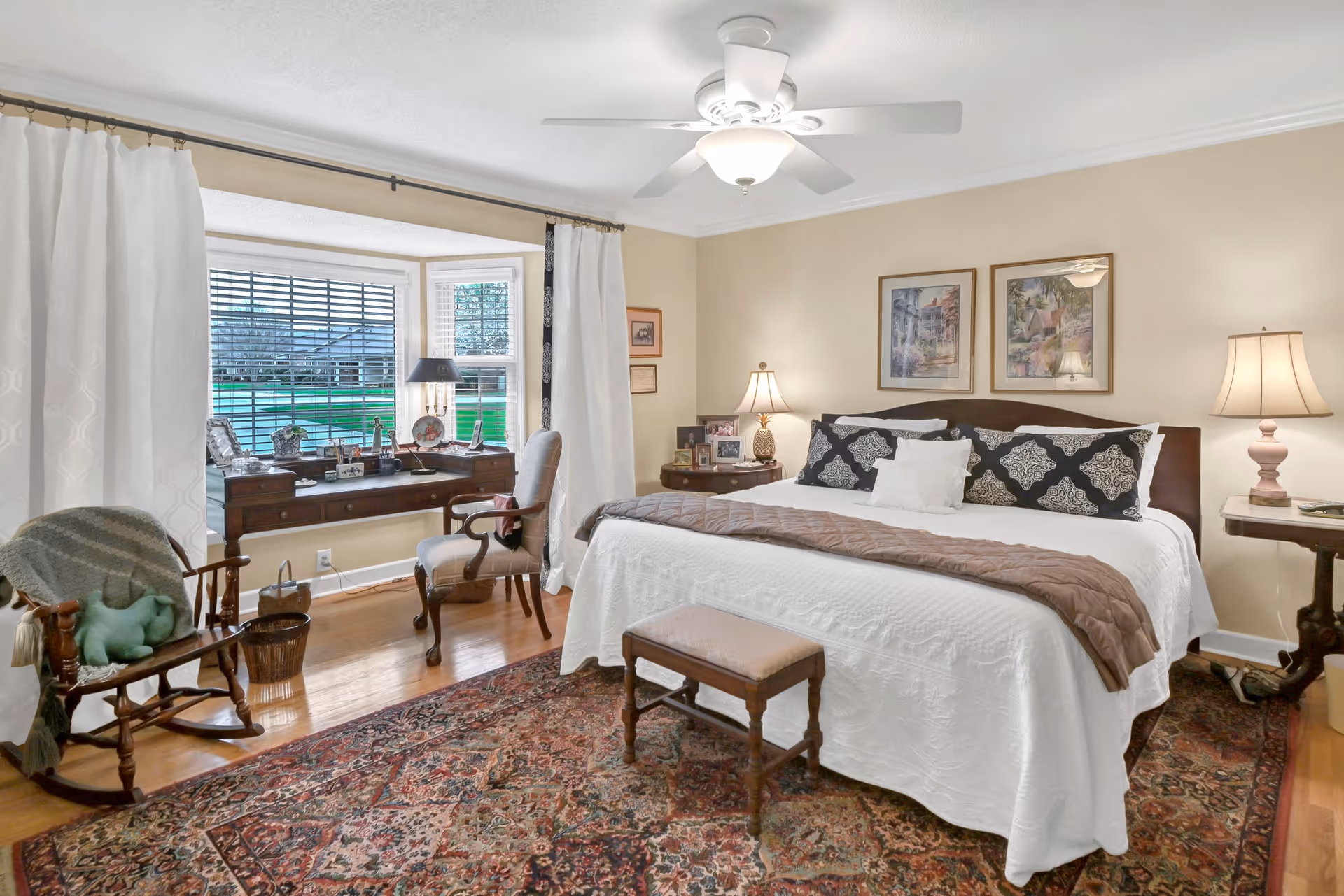 A cozy bedroom with a large bed covered in white bedding and decorative pillows. There is a small bench at the foot of the bed, two bedside tables with lamps, and framed artwork on the wall above the bed. A wooden rocking chair with a green pillow and a blanket is placed near a window with white curtains. A wooden desk with a chair is positioned in front of the window, which looks out onto a green lawn. The room has hardwood floors and a patterned area rug, with a ceiling fan overhead.