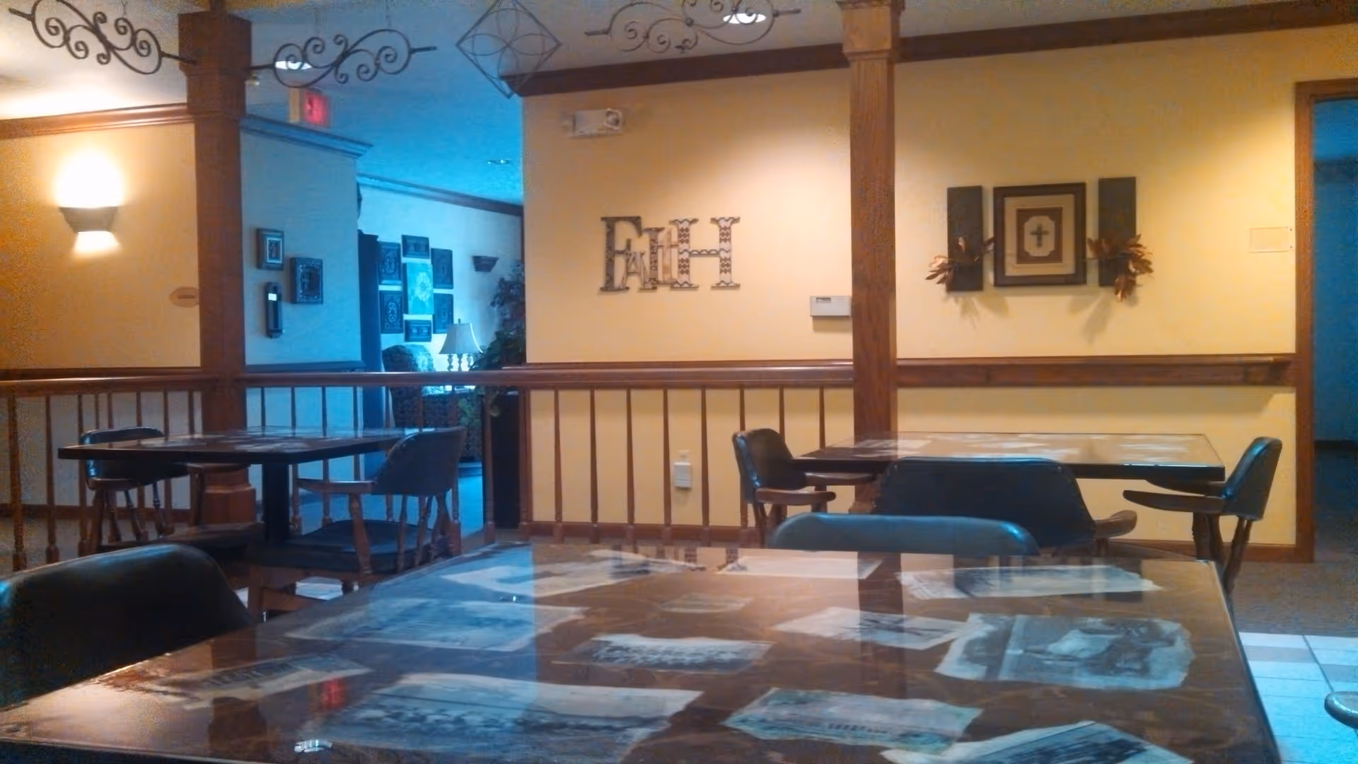 Interior view of a common area in a senior living facility with tables and chairs. The walls are painted yellow with wooden trim and decorated with wall art, including a large decorative 'Faith' sign and framed pictures. There is soft lighting from wall sconces and a lamp in the background.