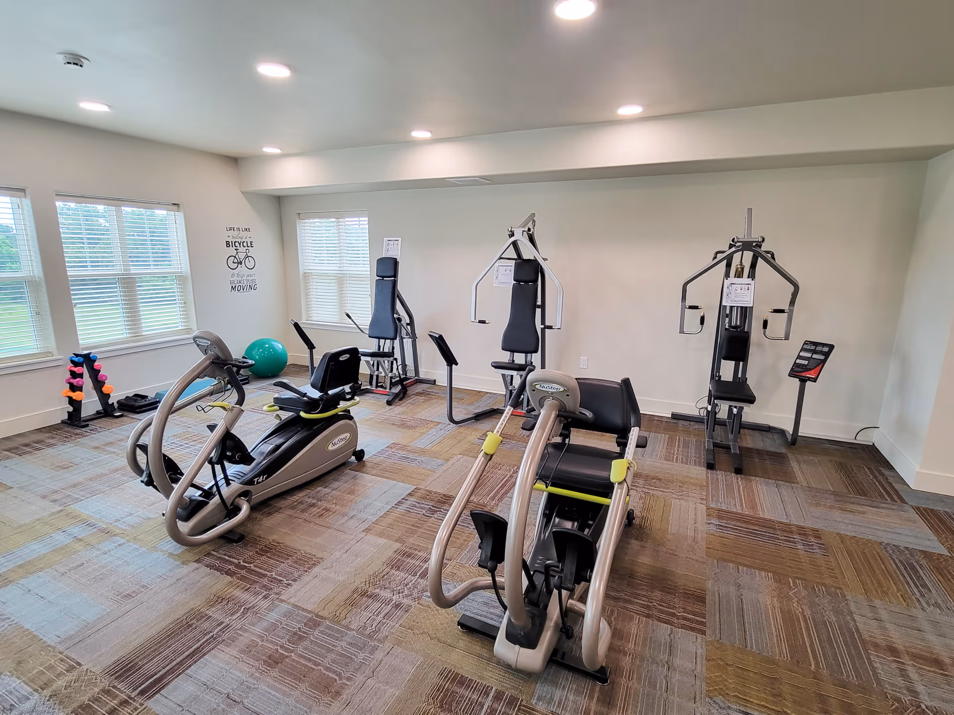 A bright exercise room with large windows letting in natural light. The room contains two NuStep recumbent cross trainers in the foreground and two multi-function weight machines against the far wall. There is a rack of colorful dumbbells and a green exercise ball near the windows. The walls are white, and the floor is covered with patterned carpet tiles. A motivational wall decal with a bicycle and the phrase 'Life is like riding a bicycle. To keep your balance, you must keep moving' is visible on the left wall.