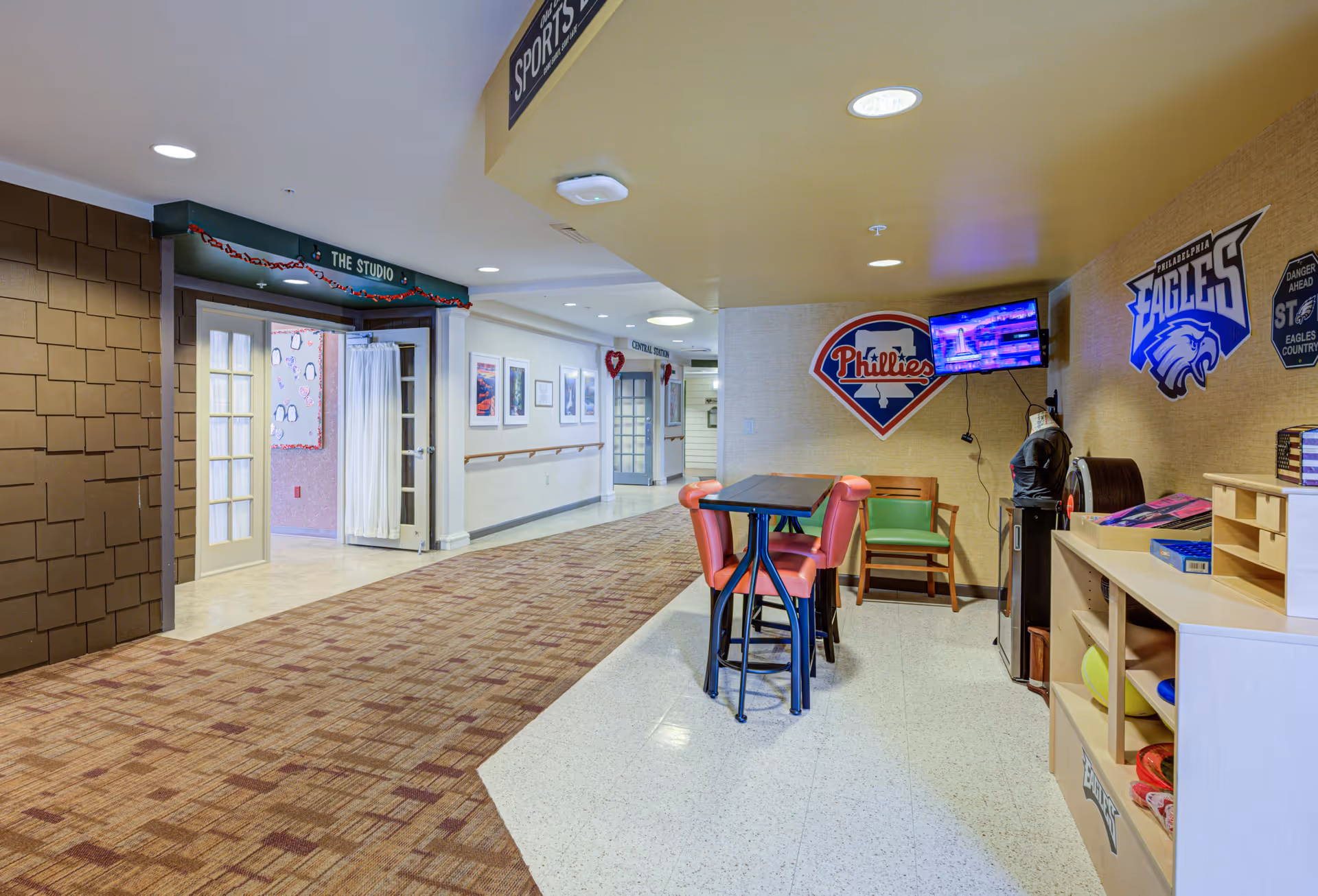 Interior hallway of Arden Courts - ProMedica Memory Care Community with a small seating area featuring a high table and chairs, sports-themed wall decorations including Philadelphia Phillies and Eagles logos, a mounted TV, and an open doorway labeled 'The Studio'.