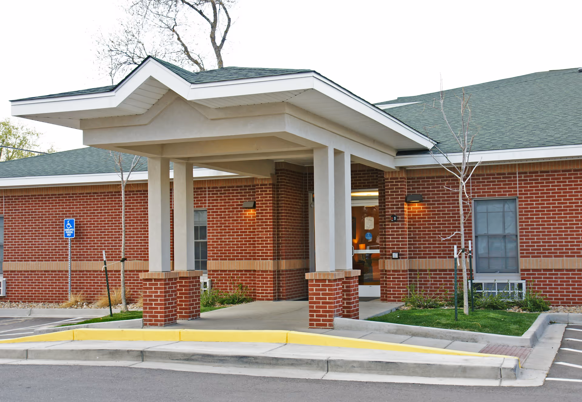 Entrance of Peaks Care Center showing a covered drop-off area with brick pillars and a green roof. There is a handicap parking sign visible near the entrance, and the building has red brick walls with windows and some small landscaping.