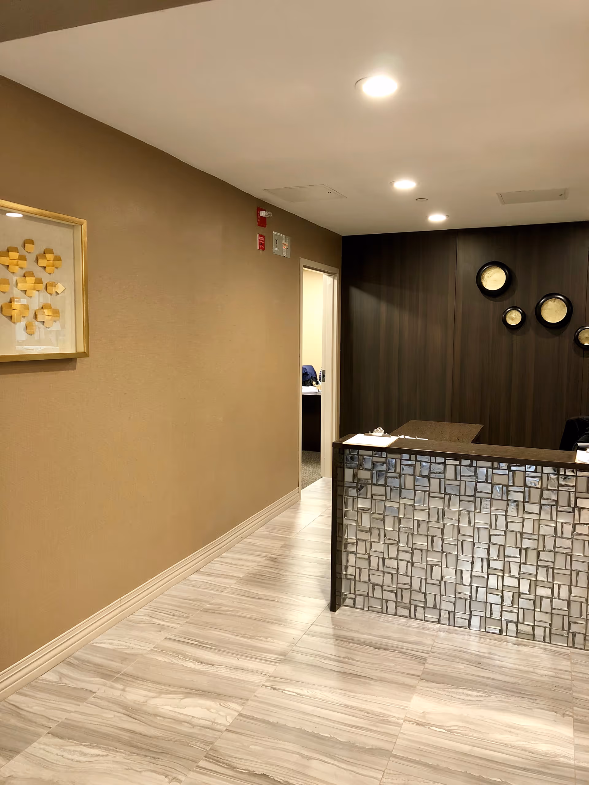 Interior view of a reception area with a tiled front desk, beige walls, a framed abstract artwork on the left wall, and a dark wood-paneled wall with circular decorative mirrors behind the desk. The floor is light-colored with a marble-like pattern, and there is an open doorway leading to another room.