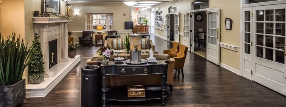 A spacious and well-lit senior living facility common area with wooden flooring, comfortable seating including armchairs and sofas, a fireplace on the left side, decorative plants, and framed pictures on the walls. There are multiple doorways with glass-paneled doors along the right side and a hallway leading further into the facility.