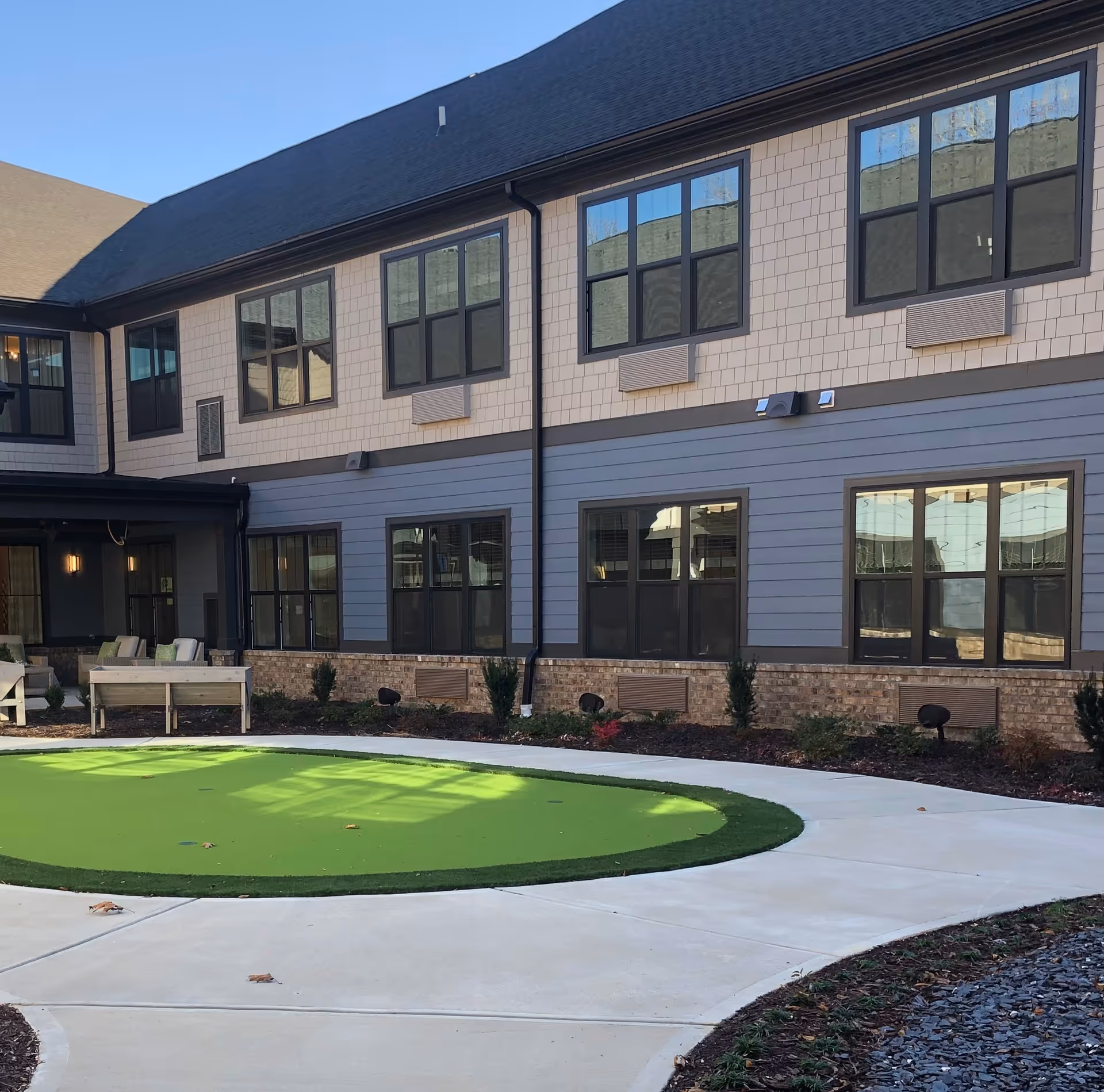 Outdoor courtyard area of a senior living facility with a circular green putting green surrounded by a concrete walkway. The building has two stories with multiple windows and a mix of brick, blue siding, and beige shingles on the exterior walls. There are some small shrubs and plants along the building's foundation and a few benches with cushions near the left side.