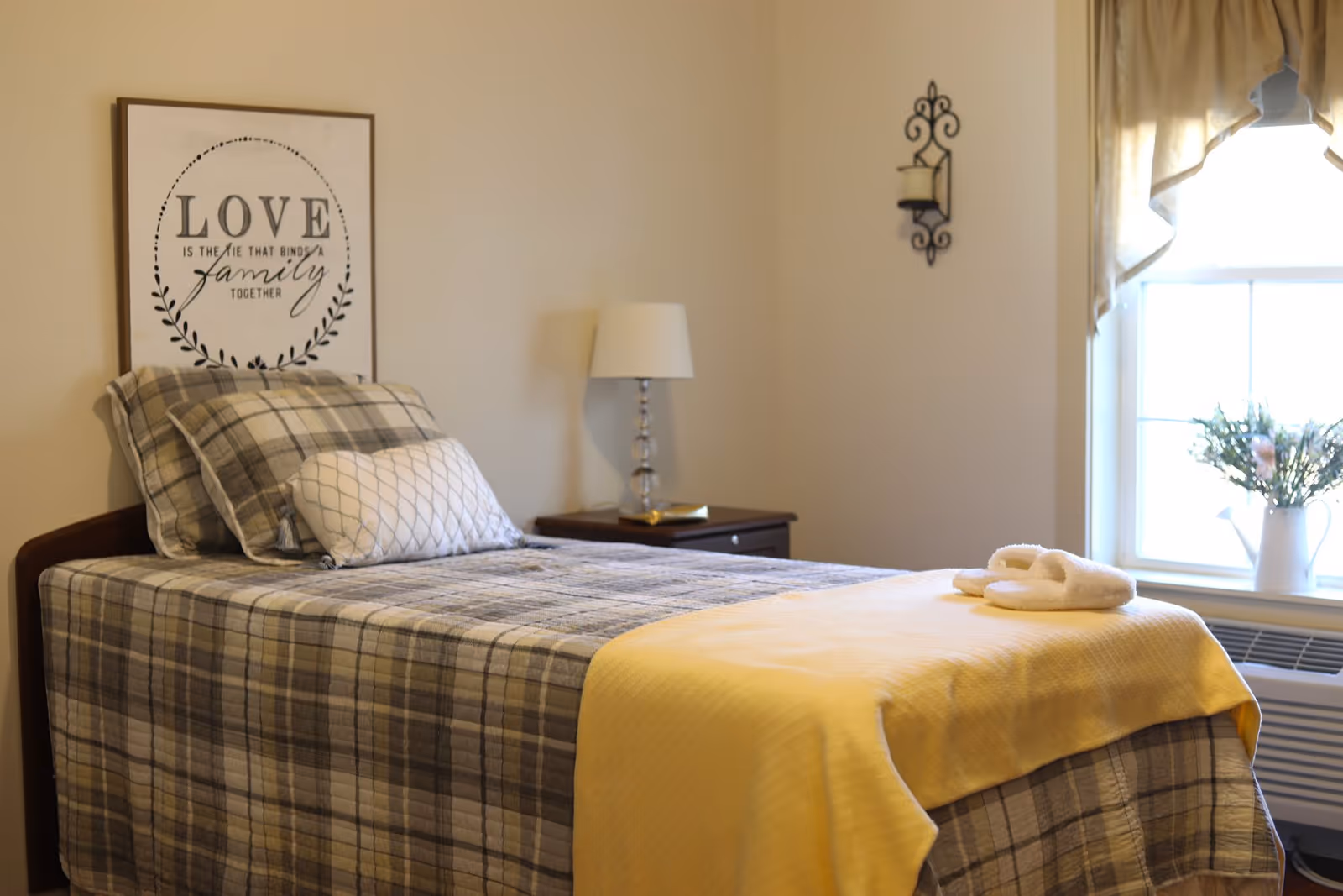 A neatly made single bed with plaid bedding and a yellow blanket at the foot. There are two pillows on the bed, one plaid and one white with a geometric pattern. A pair of white slippers is placed on the yellow blanket. Behind the bed, there is a framed wall art with the quote 'LOVE is the tie that binds a family together.' A bedside table with a lamp and a book is next to the bed. A window with beige curtains and a white vase with flowers is on the right side of the room.
