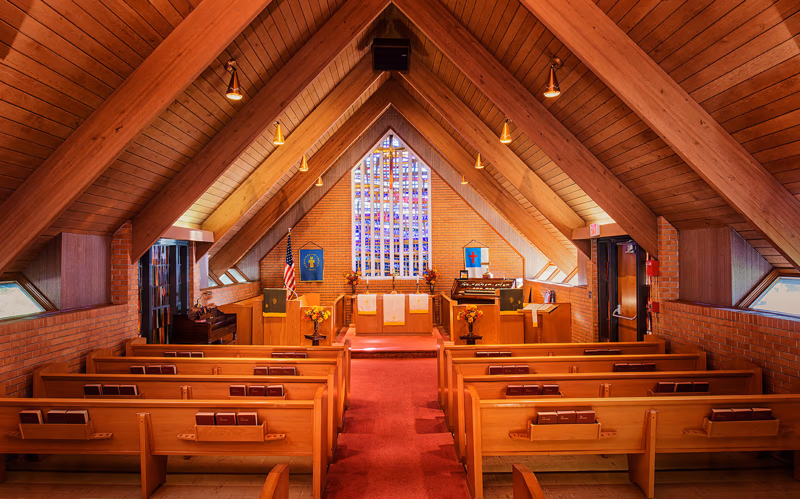 Interior view of a chapel with wooden pews arranged in rows facing an altar. The chapel has a high wooden ceiling with exposed beams and a large stained glass window behind the altar. There are floral arrangements on either side of the altar, an American flag, and religious banners on the walls. The floor is carpeted in red.