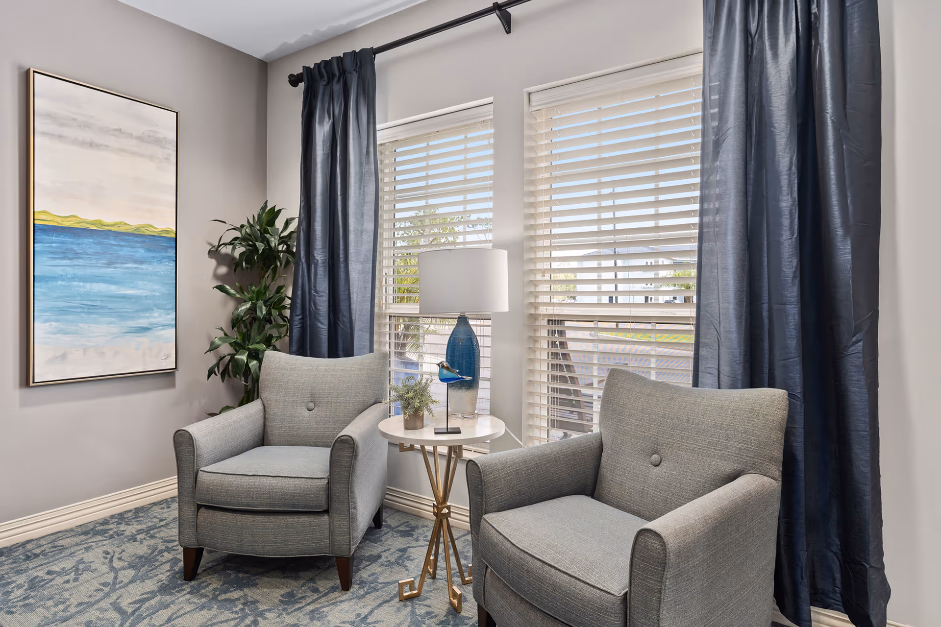 Two gray armchairs flank a small round table with a lamp between windows with blinds and dark curtains, next to a plant and a coastal painting.