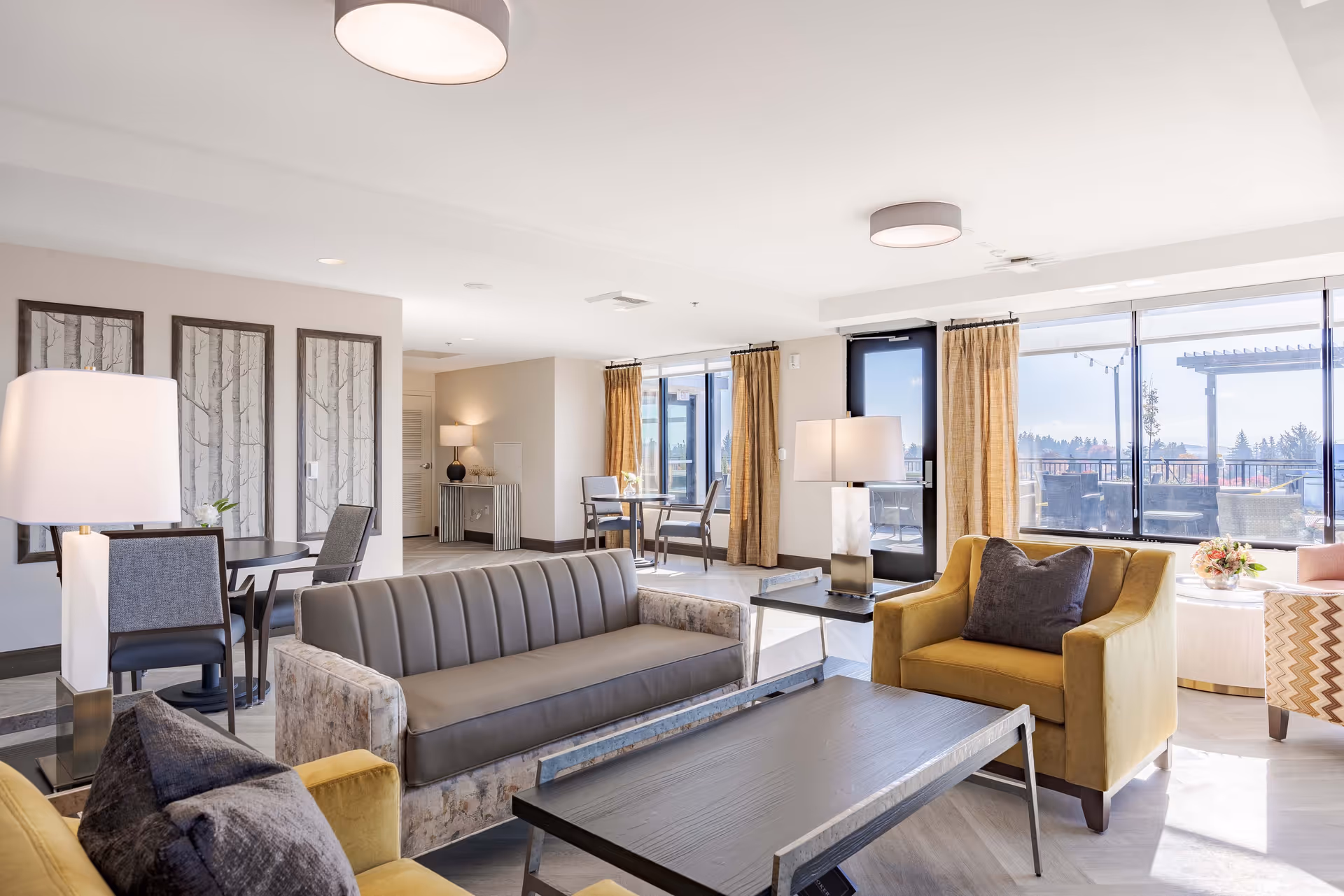 Sunlit modern lounge with sofas, armchairs, a coffee table, and large windows overlooking an outdoor terrace.