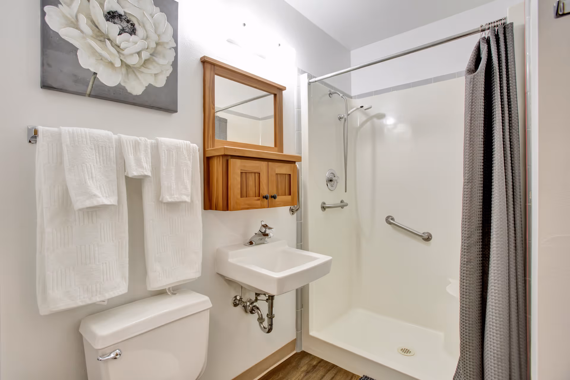 Small bright bathroom featuring a walk-in shower with grab bars, a wall-mounted sink, toilet, towel rack, and wooden medicine cabinet.