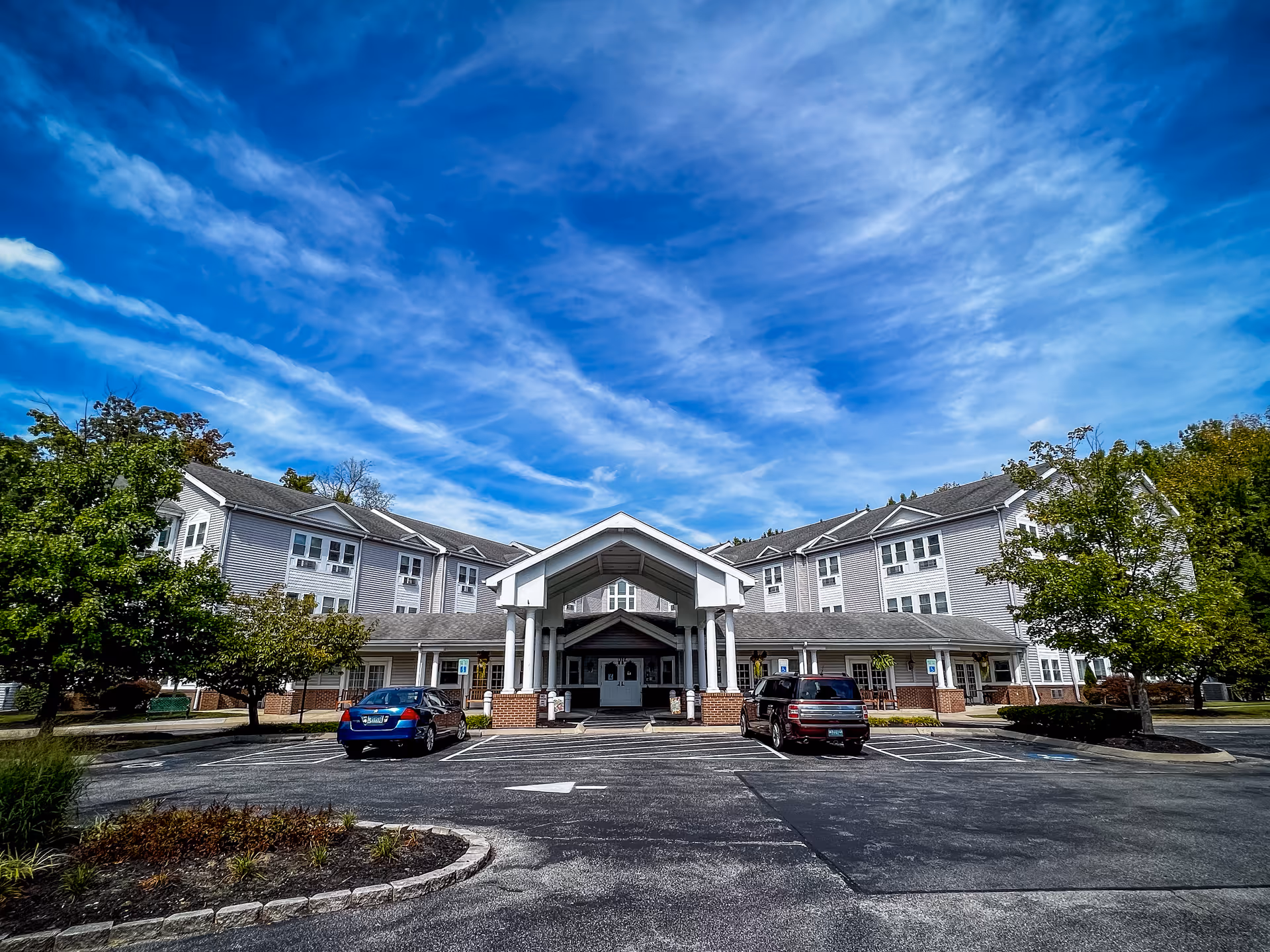 Front exterior view of Inspirations Bayside Assisted Living & Memory Care facility with a covered entrance, two parked cars, trees on both sides, and a bright blue sky with scattered clouds.