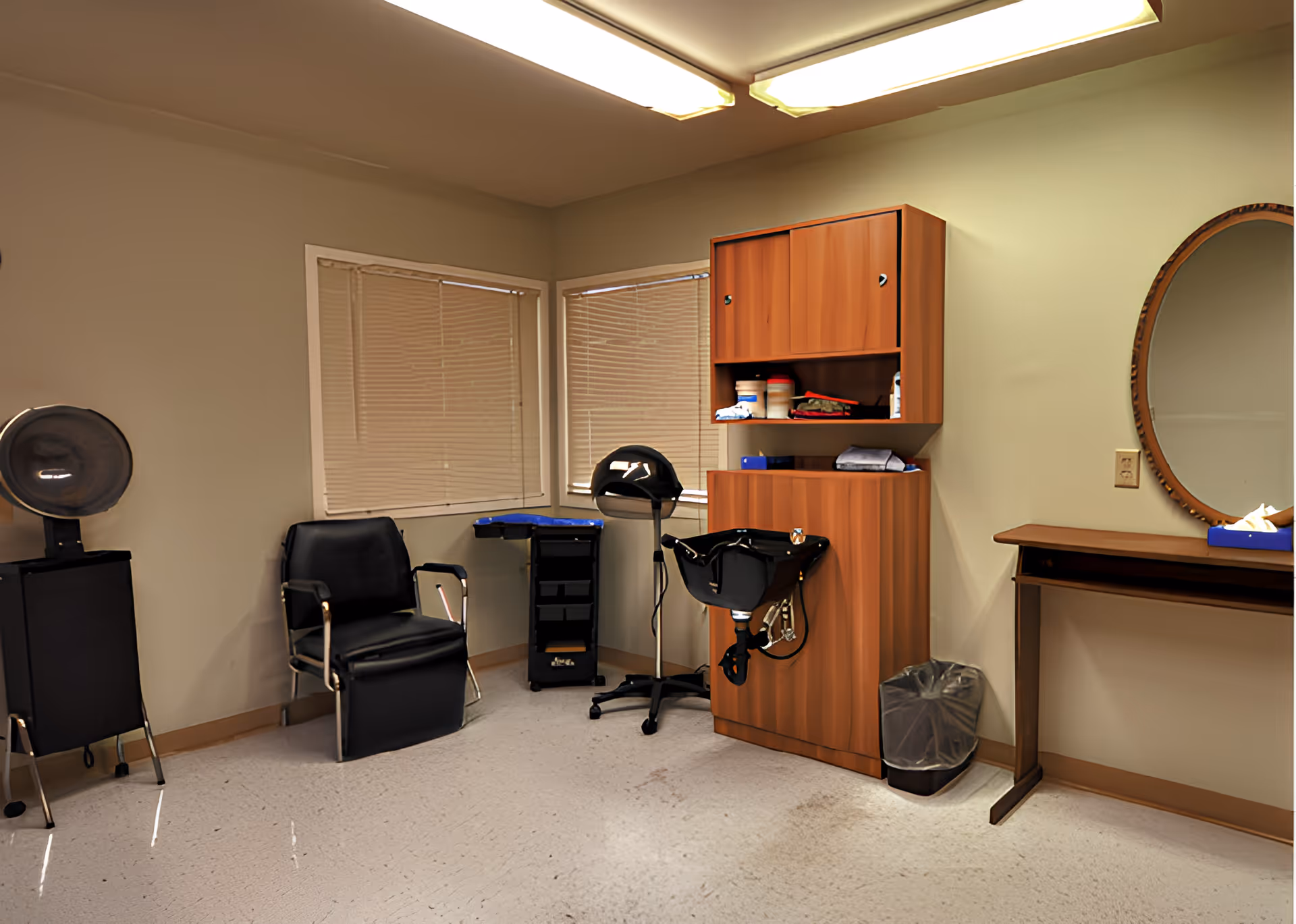 Interior of a hair salon area with a black salon chair, hair dryer, wooden cabinets with hair products, a mirror on the wall, and a small table with a tissue box.
