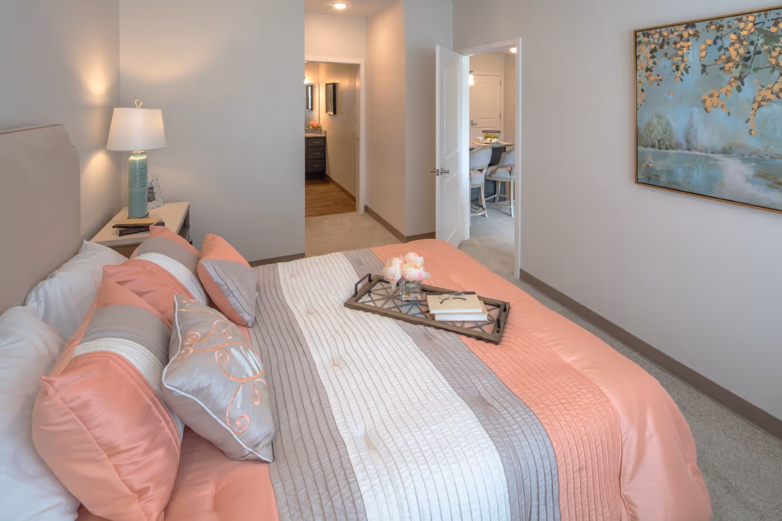 A neatly made bed with a pink, gray, and white striped comforter and multiple pillows in a bedroom. A bedside table with a lamp and picture frame is next to the bed. A tray with flowers and a book is placed on the bed. The room has light-colored walls, a painting on the wall, and an open door leading to a hallway and another room with a dining table and chairs.