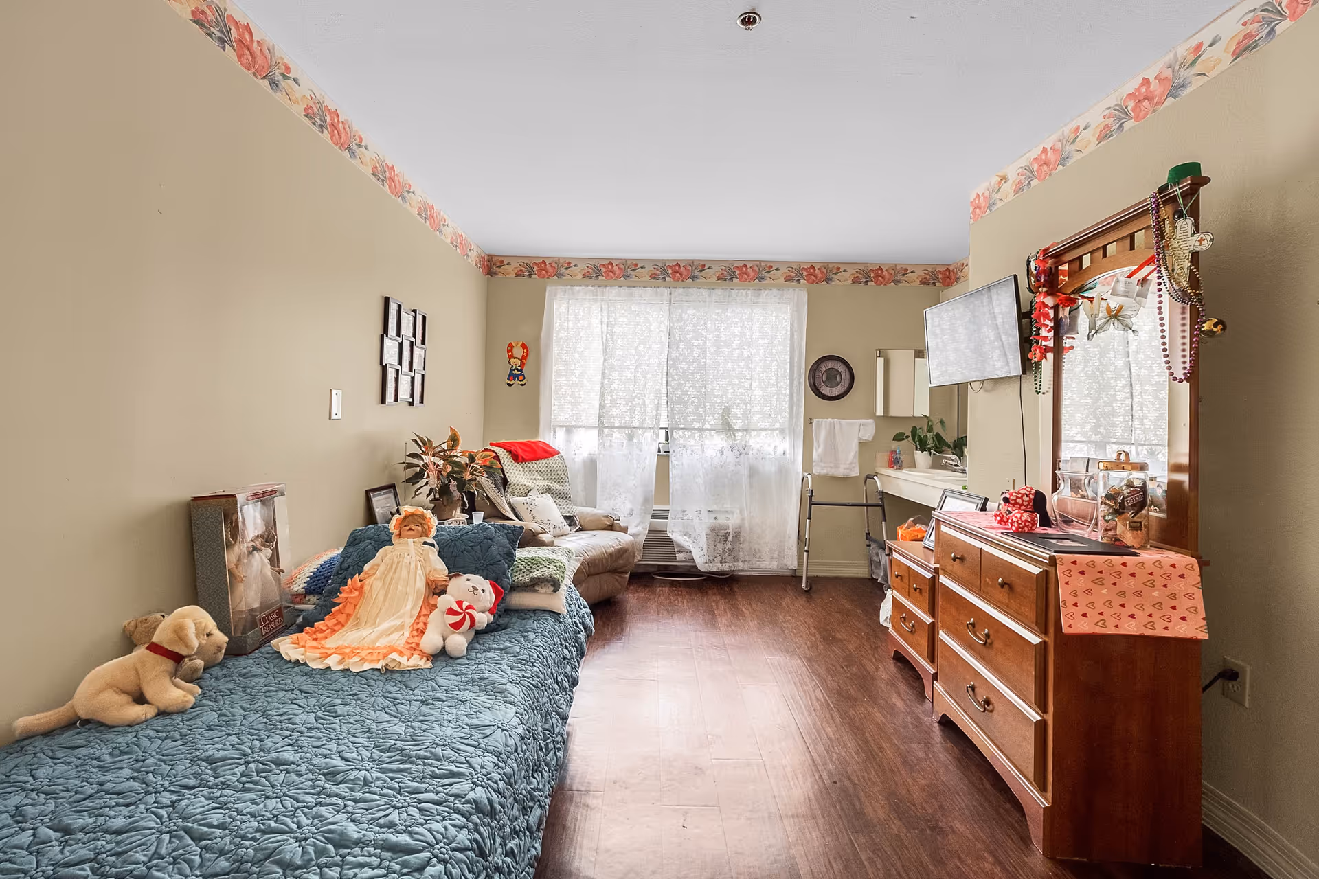 A cozy bedroom in a senior living facility with a single bed covered in a blue quilt and decorated with dolls and stuffed animals. There is a comfortable armchair near a window with sheer white curtains, a wooden dresser with a mirror adorned with beads and decorations, a wall-mounted TV, and a walker near a small sink area. The walls are beige with floral border wallpaper near the ceiling.