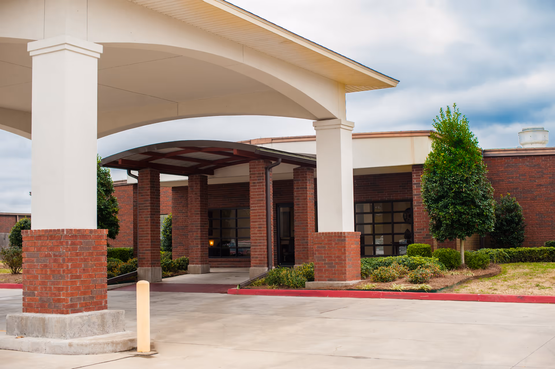 Entrance of a brick building with a covered drop-off area supported by white columns, surrounded by neatly trimmed bushes and small trees under a cloudy sky.