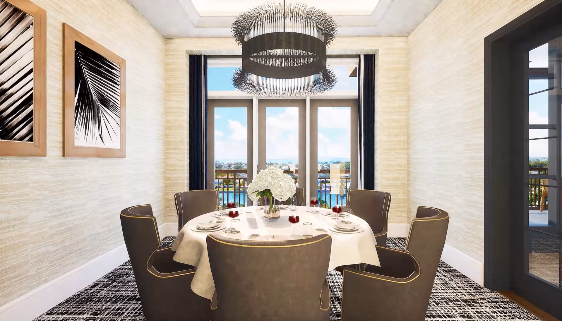 A bright dining room with a round table covered with a white tablecloth, set with plates, glasses of red wine, and a centerpiece of white flowers. Six upholstered chairs surround the table. The room features large windows with a view of a balcony and water beyond, two framed black and white palm leaf prints on the wall, and a modern chandelier hanging from the ceiling.