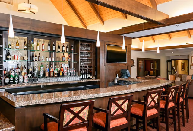 Interior view of a bar area with a curved granite countertop and several wooden bar stools with red cushioned seats. Behind the bar are shelves stocked with various bottles of liquor and wine glasses. The ceiling has wooden beams and pendant lights hanging above the bar. A flat screen TV is mounted on the wall behind the bar.
