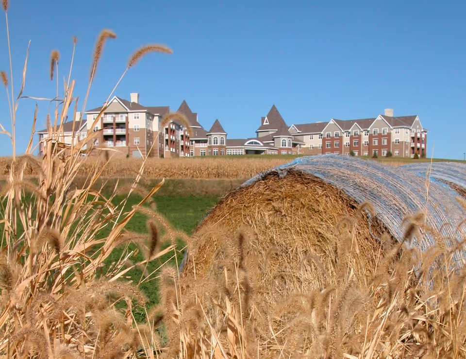 A large senior living facility building with multiple peaked roofs and balconies, situated on a hill behind a field with tall dry grasses and a large round hay bale wrapped in netting under a clear blue sky.