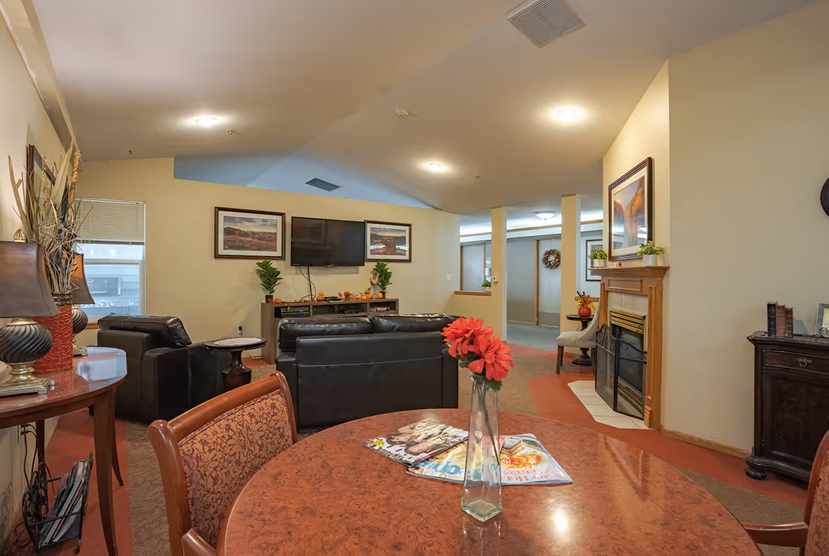 Communal living room with leather sofas facing a wall-mounted TV, a fireplace, and a table in the foreground with a vase of red flowers and magazines.