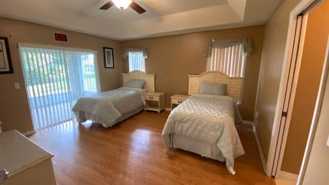 A bedroom in a senior living facility with two single beds, each with a white headboard and light-colored bedding. There are two nightstands between the beds, a dresser in the foreground, and windows with vertical blinds and valances. The room has wooden flooring and beige walls.