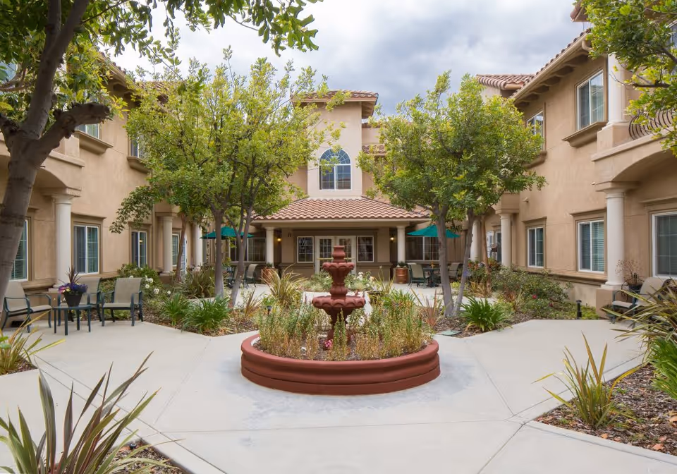 Outdoor courtyard area of a senior living facility with a central red fountain surrounded by plants and trees. The courtyard is paved with concrete walkways and has seating areas with chairs and tables under green umbrellas. The building surrounding the courtyard has beige stucco walls, multiple windows, and a tiled roof.