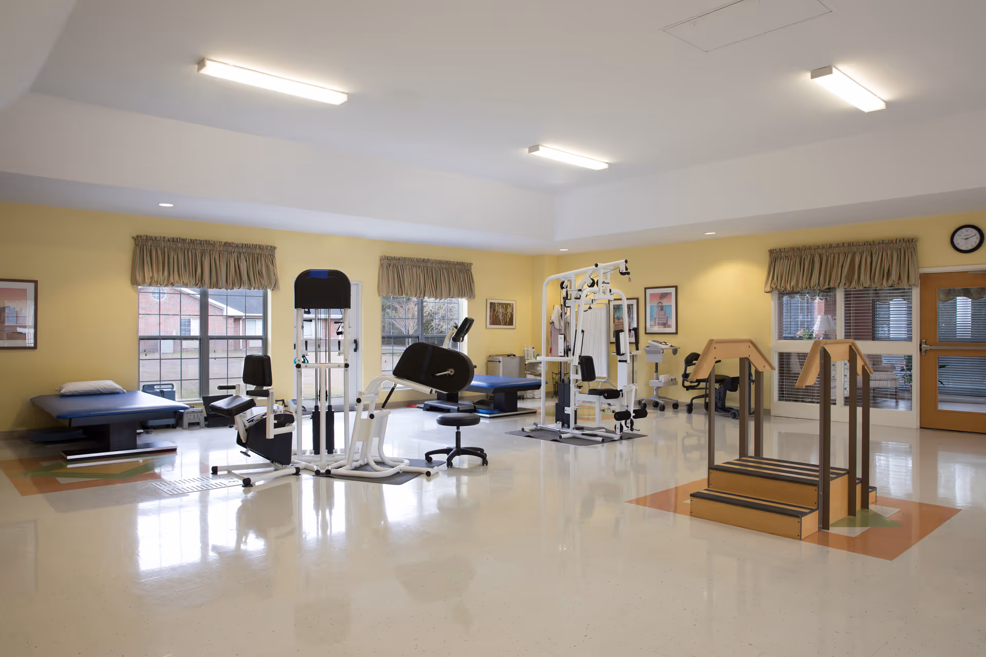 A spacious physical therapy or rehabilitation room with exercise equipment including a stationary bike, weight machines, therapy tables, and a small set of stairs with handrails. The room has large windows with beige curtains, yellow walls, and a polished floor. There are framed pictures on the walls and a clock above a door.