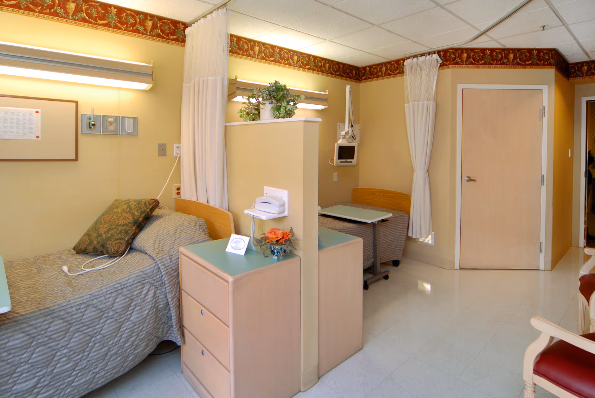 A clean and well-lit room with two hospital-style beds separated by a partial divider with a small cabinet. Each bed has a reading light above it, a pillow, and a bedside table. There are curtains that can be drawn for privacy, a door in the background, and a chair with red upholstery partially visible on the right.