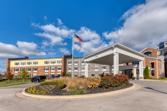 Exterior view of a senior living facility building with a covered entrance, landscaped roundabout with flowers and bushes, an American flag on a flagpole, and a partly cloudy blue sky.