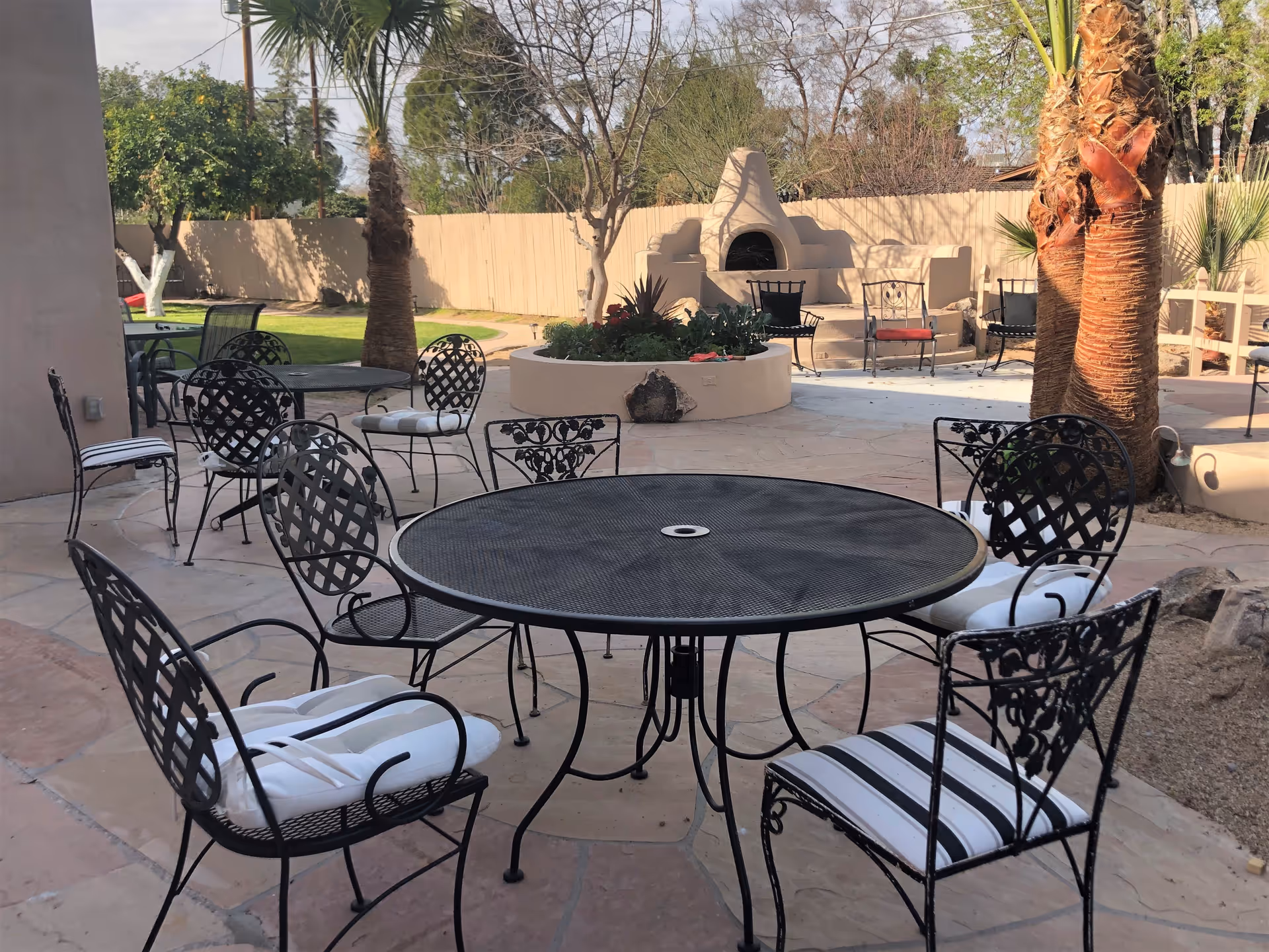 Outdoor patio area with several black metal tables and chairs with cushions, surrounded by palm trees and other greenery. In the background, there is a beige outdoor fireplace and a circular planter with plants.