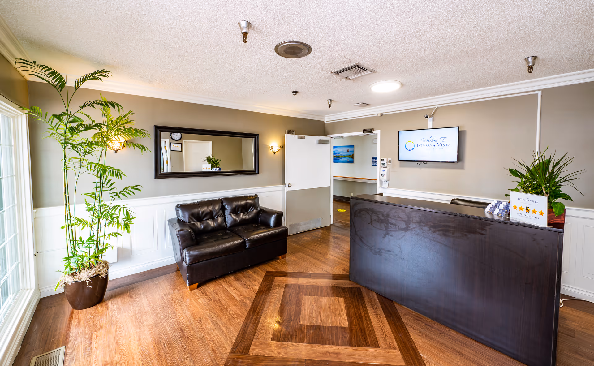 Reception area of Pomona Vista Care Center with a dark wooden front desk, a black leather couch, a large mirror on the wall, potted plants, and a wall-mounted TV displaying the facility's welcome message.