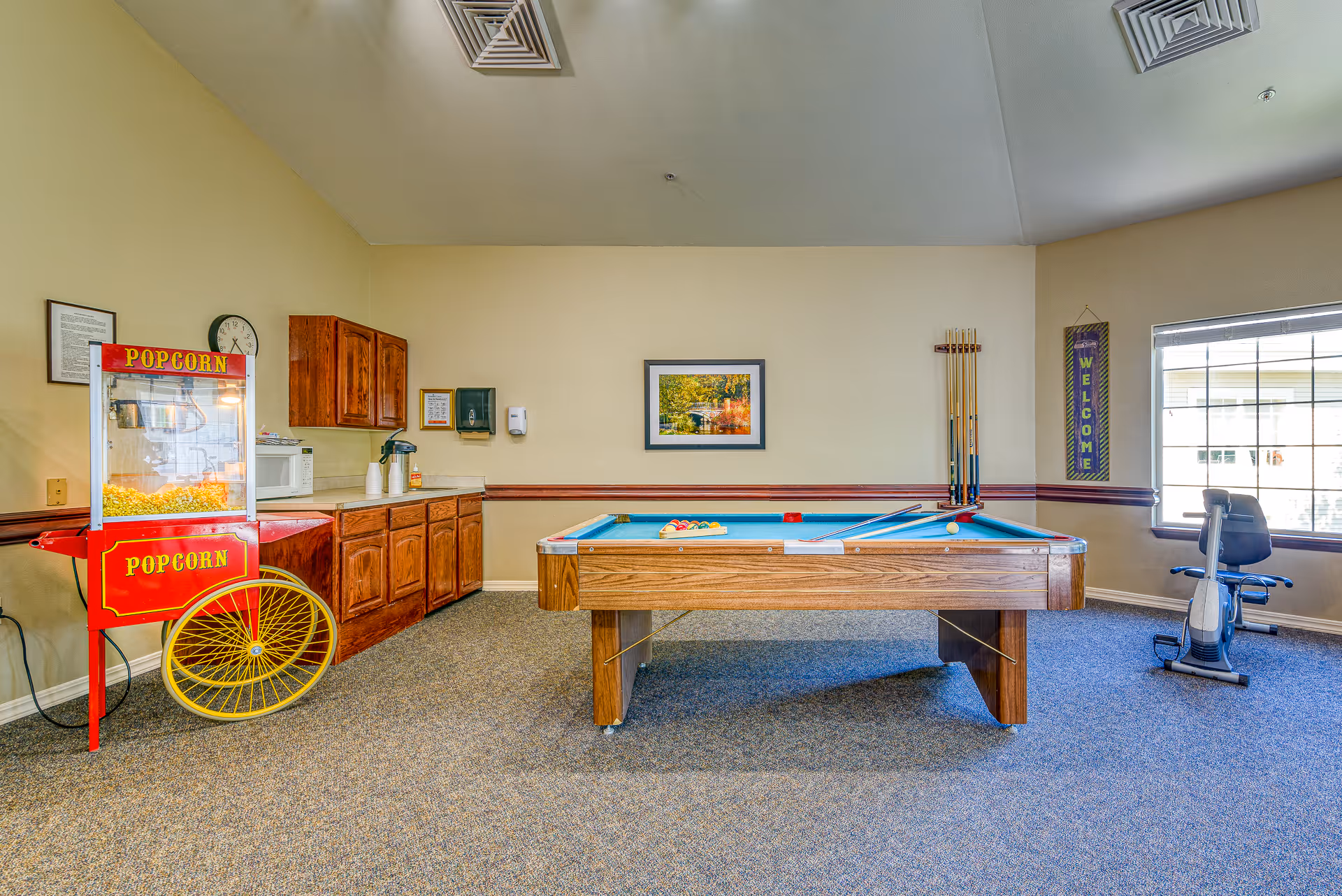 A bright recreation room with a pool table at center, a red popcorn cart, kitchenette cabinets, and an exercise bike by the window.