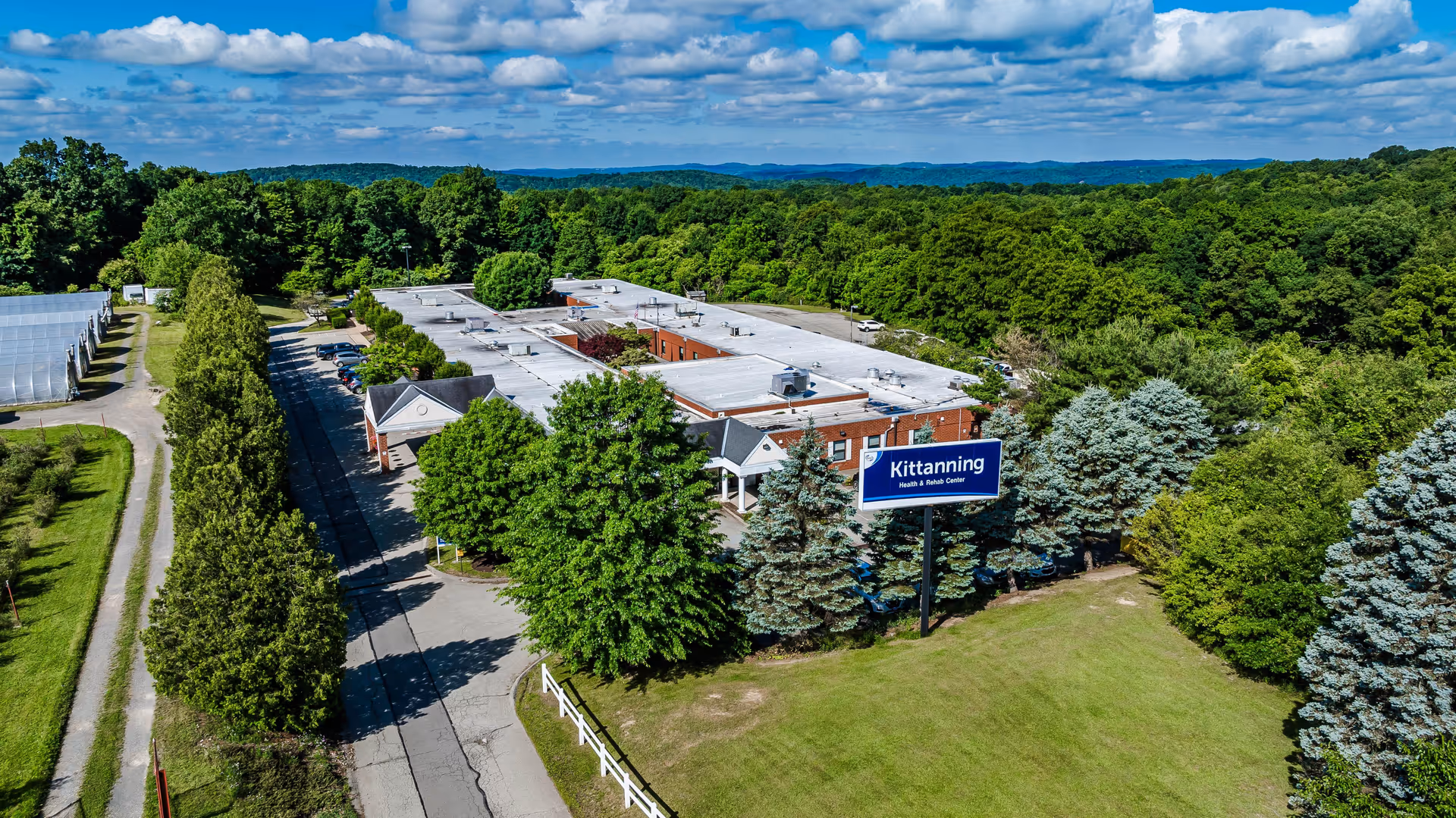 Aerial view of Kittanning Health & Rehab Center surrounded by trees and greenery under a partly cloudy sky. The building has a flat roof and a large sign in front displaying the facility's name. There is a driveway lined with trees leading to the entrance.