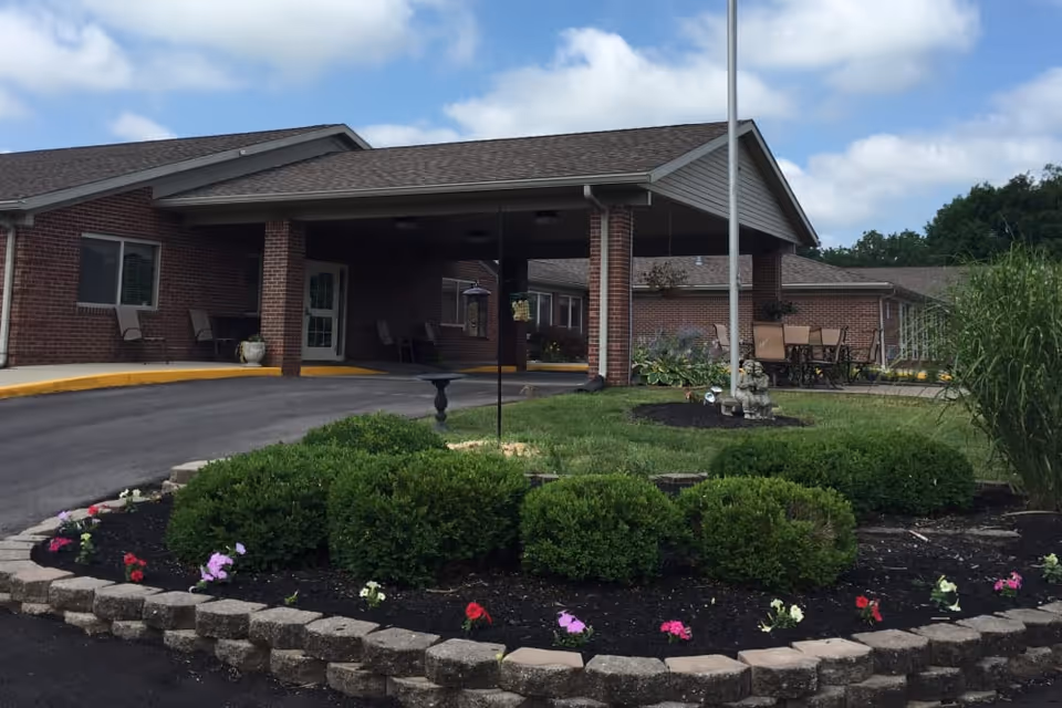 Front exterior view of a single-story brick building with a covered entrance and a circular driveway. There is a landscaped garden with green bushes and colorful flowers in front of the building, along with a flagpole and outdoor seating area with chairs and tables. The sky is partly cloudy.