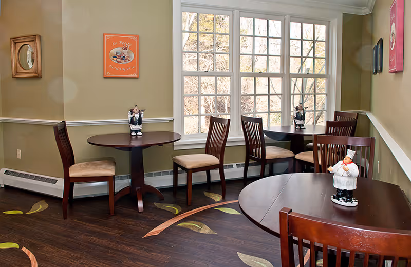 A small dining area with three round wooden tables and six wooden chairs with beige cushions. Each table has a decorative chef figurine on it. The room has light green walls, a large window letting in natural light, and a dark wooden floor with a leaf pattern. There are two framed pictures on the walls.