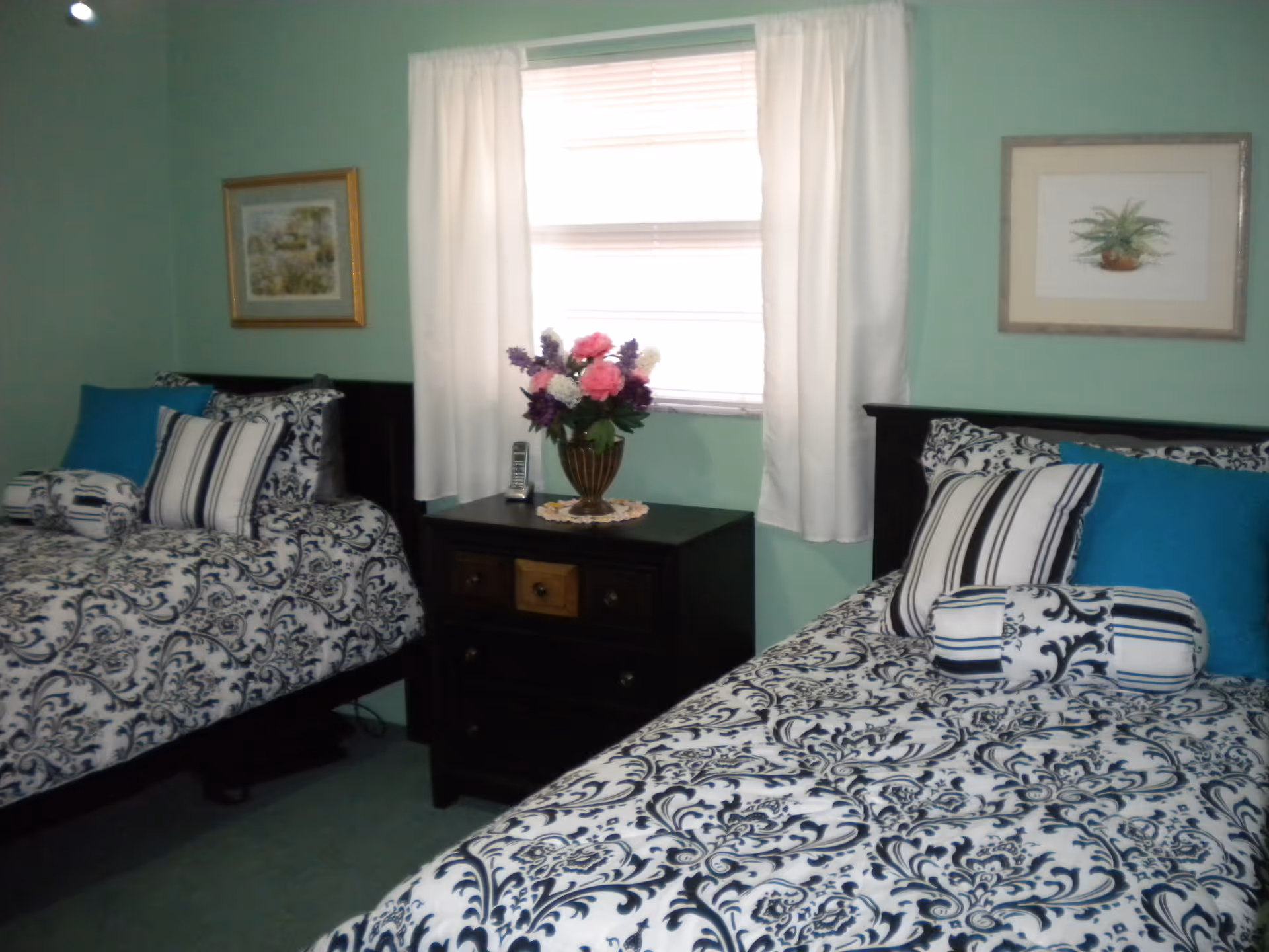Two twin beds with black headboards and blue-and-white patterned bedding flank a central nightstand topped with a vase of flowers beneath a curtained window.