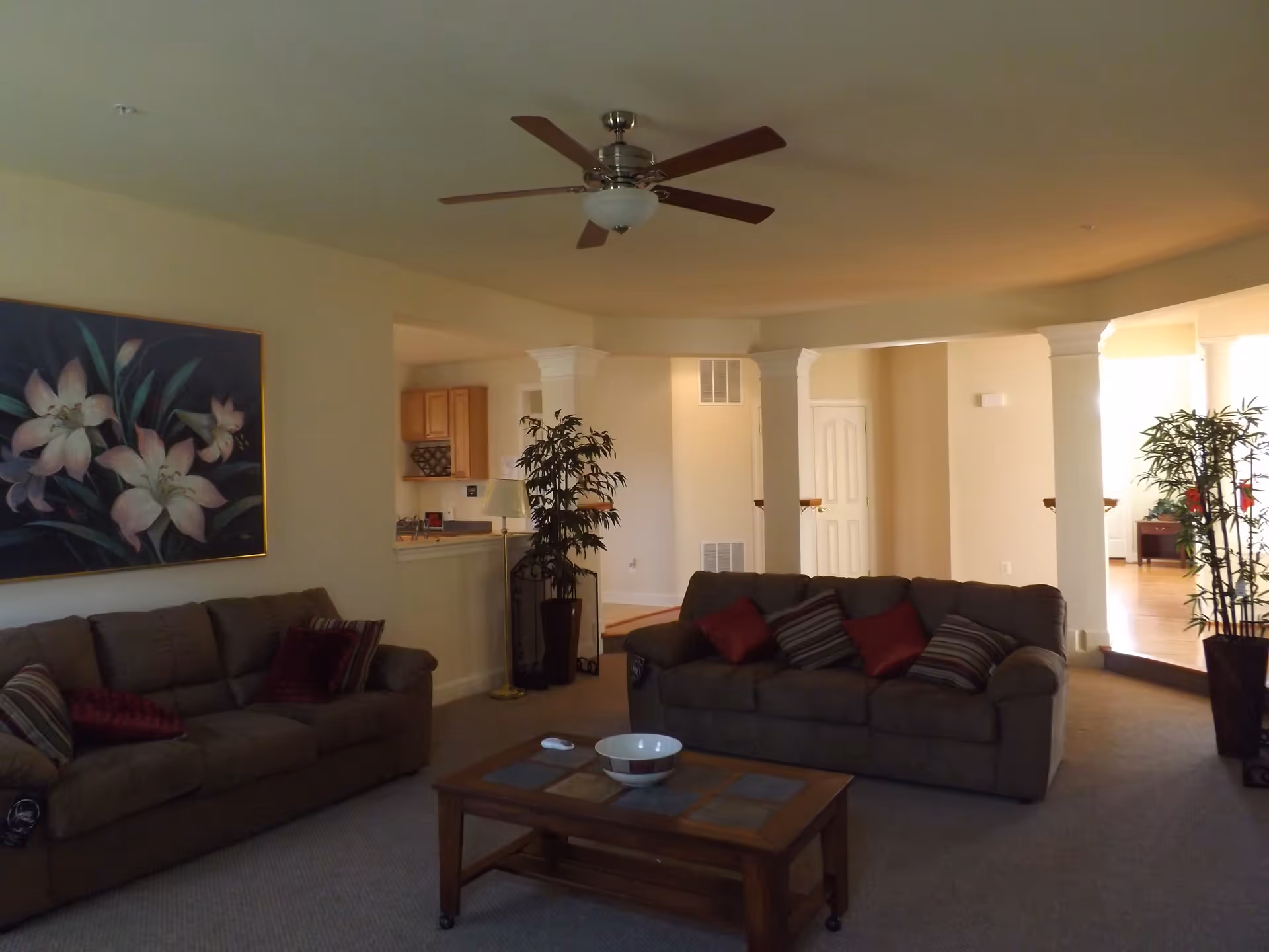A living room with two brown sofas adorned with red and striped pillows, a wooden coffee table with a white bowl on top, a large floral painting on the wall, a ceiling fan, and potted plants near white columns separating the living area from an adjacent space.