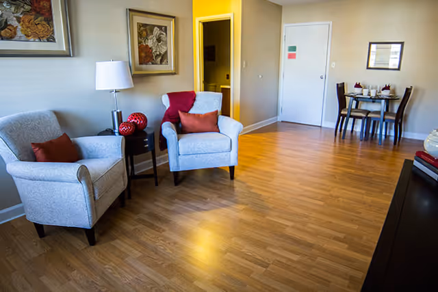 A cozy living room area with two light blue upholstered armchairs, each with a red throw pillow. Between the chairs is a small round black side table with a white lamp and decorative red spheres. The room has wooden flooring and beige walls adorned with framed floral artwork. In the background, there is a small dining area with a table set for four and a white door with colorful notes attached.