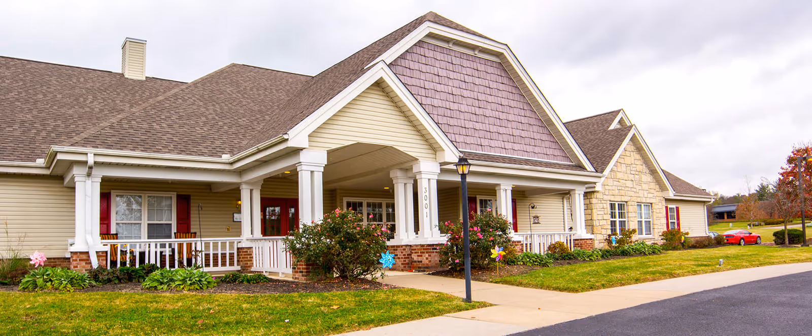 Exterior view of a single-story senior living facility building with beige siding, white columns, and a covered entrance. There are bushes and flowers planted along the front, a sidewalk leading to the entrance, and a red car parked on the right side. The sky is overcast.