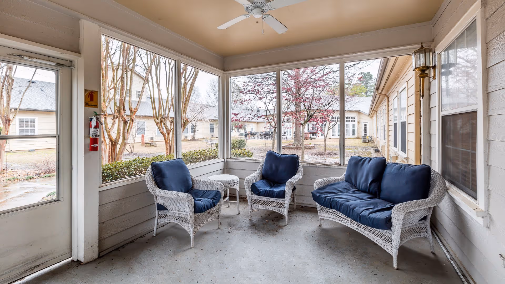 A screened-in porch area with white wicker furniture including two armchairs and a loveseat, all with dark blue cushions. There is a small round wicker table between the chairs. The porch overlooks a courtyard with trees and buildings in the background.