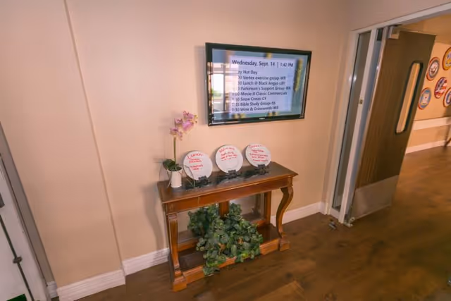 Interior hallway of Lexington Assisted Living with a wooden table against a beige wall. On the table are three white plates with red writing, a small potted orchid, and some green foliage underneath. Above the table, a wall-mounted screen displays a schedule of activities for Wednesday, Sept. 14. To the right, a door with a window leads to another room with framed pictures on the wall.