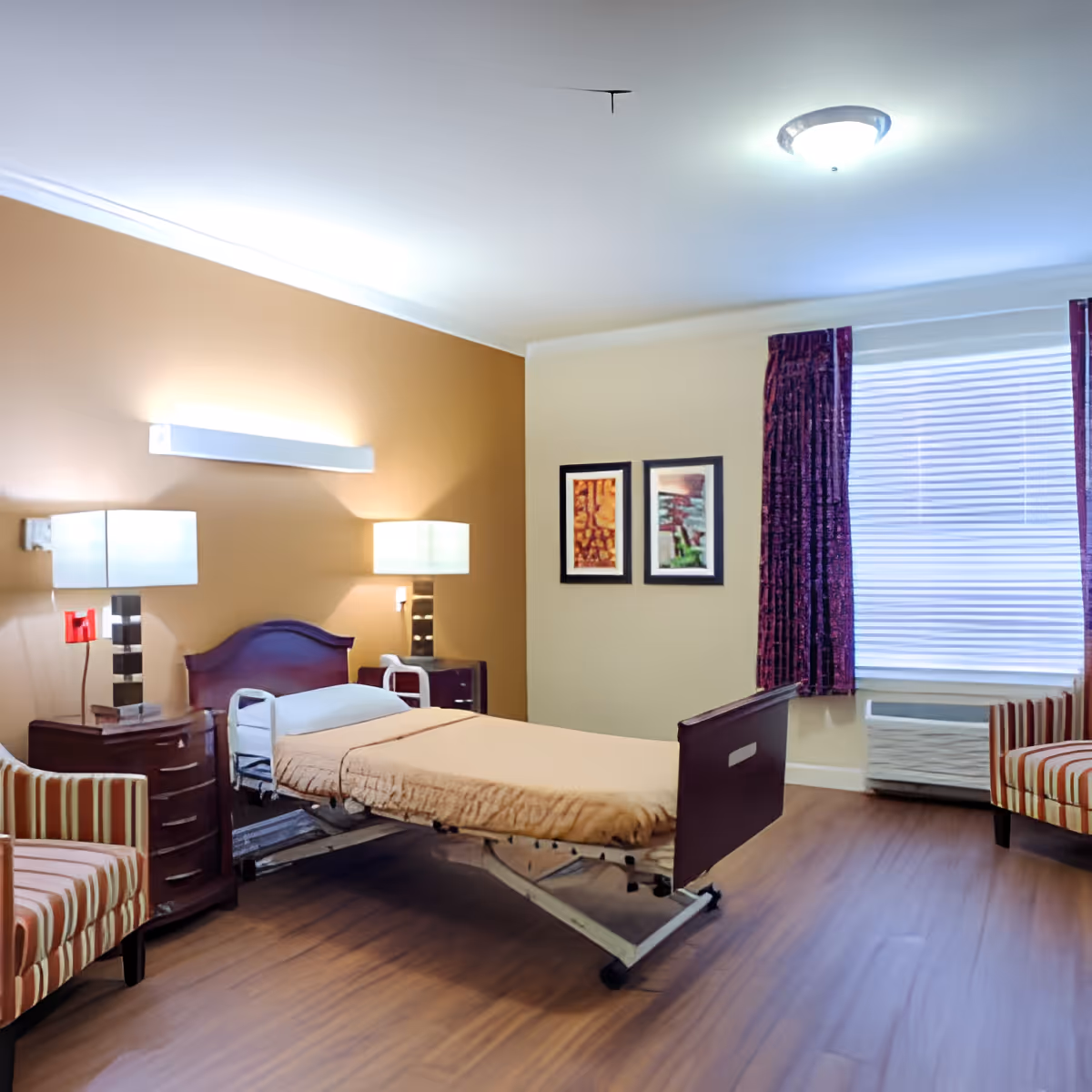 A senior living facility bedroom with a hospital-style adjustable bed covered with a beige blanket. The room has two striped armchairs, two bedside tables with modern lamps, two framed pictures on the wall, a window with closed blinds and purple curtains, and wood flooring.