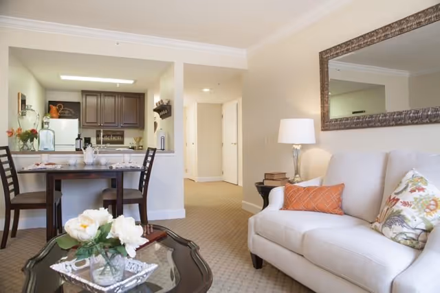 Bright living room with a beige sofa, coffee table with flowers, a dining table, and an open kitchen visible in the background.