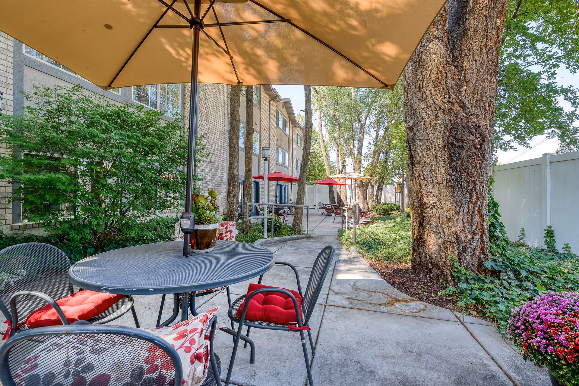 Outdoor patio area at The Laurel at Millcreek featuring a round metal table with red and floral cushions on black metal chairs, shaded by a large beige umbrella. The patio is surrounded by trees, green bushes, and a white fence, with additional tables and umbrellas visible in the background along a concrete pathway.