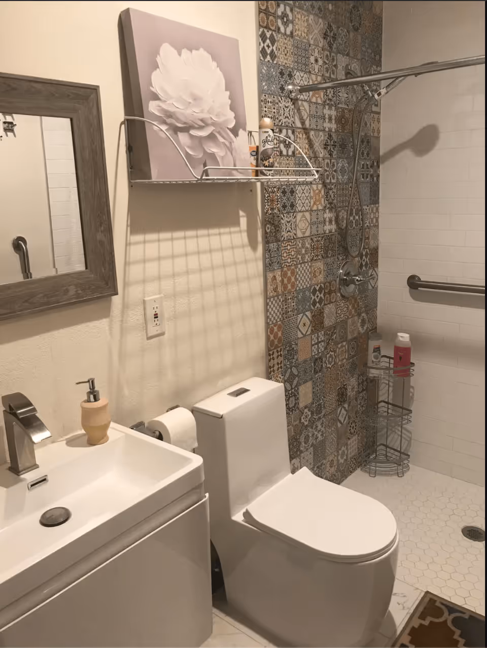 A bathroom with a white toilet and sink. The wall behind the toilet features a decorative tile pattern in various colors and designs. There is a shower area with a handheld showerhead and a grab bar. A metal shelf above the toilet holds a can of air freshener and other items. A mirror with a wooden frame hangs above the sink, and a soap dispenser is placed on the sink.