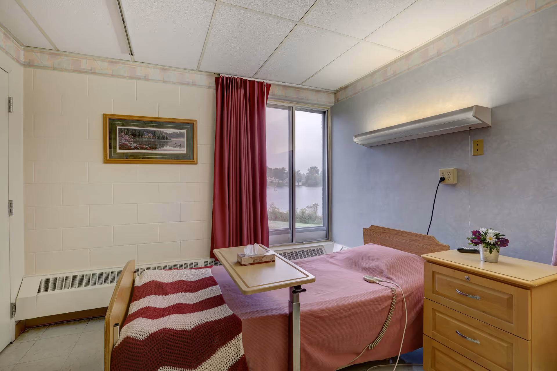 Small resident bedroom with a single bed, overbed table, wooden nightstand, and a window showing a lakeside view.