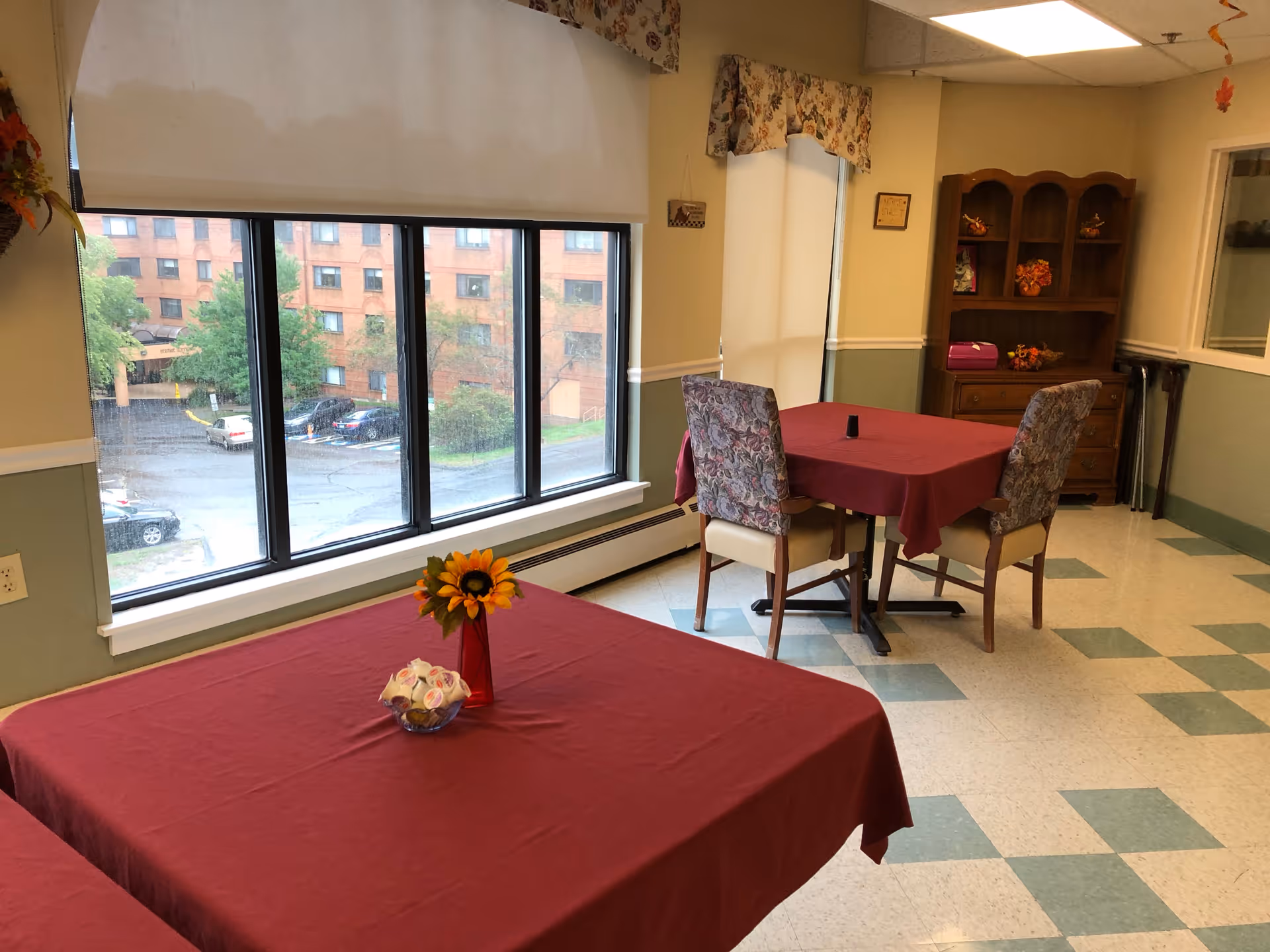 A dining area with two tables covered in red tablecloths, each with floral centerpieces. Two chairs with floral upholstery are positioned at one table. Large windows show a view of a parking lot and a brick building outside. The room has a wooden cabinet with decorative items and a tiled floor with a green and white pattern.