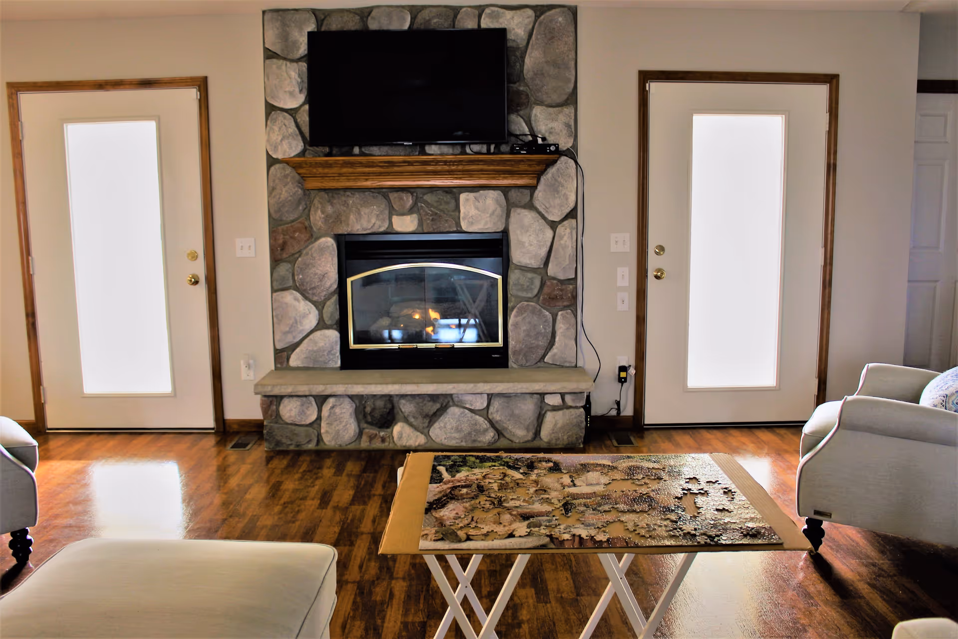 A cozy living room with a stone fireplace and a mounted flat-screen TV above it. There are two white doors with glass panels on either side of the fireplace. In front of the fireplace is a folding table with a partially completed jigsaw puzzle. The room has wooden flooring and light-colored walls, with a white armchair visible on the right side.