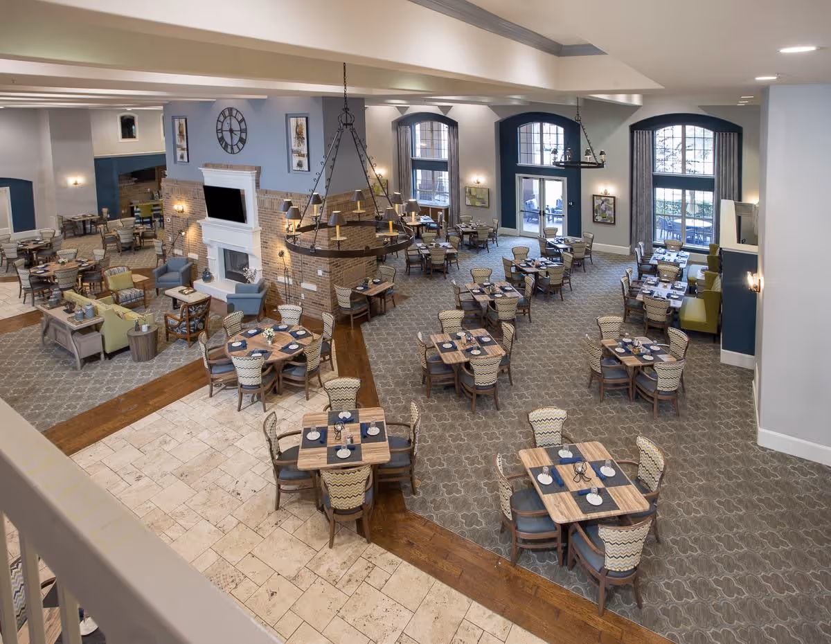 Spacious dining room in a senior living facility with multiple wooden tables and chairs arranged neatly. The room features a large fireplace with a TV mounted above it, large windows allowing natural light, and elegant chandeliers hanging from the ceiling. There are also comfortable seating areas with armchairs and sofas near the fireplace.