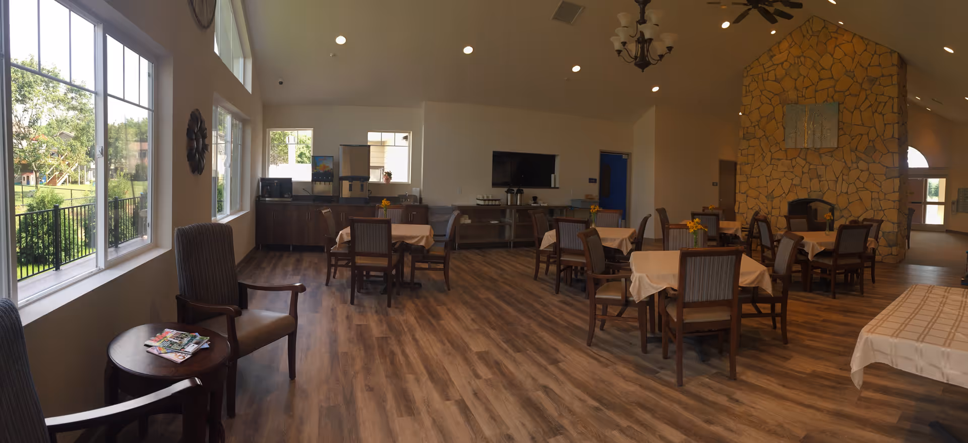 A spacious dining room in a senior living facility with several tables covered with beige tablecloths and surrounded by wooden chairs. There is a stone fireplace on the right wall with a decorative painting above it. Large windows on the left side let in natural light and offer a view of greenery outside. The floor is wood-patterned, and there are ceiling lights and a chandelier providing additional lighting. A small table with magazines and two armchairs are near the windows.