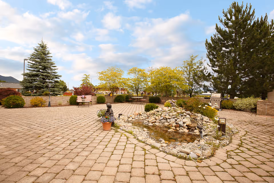 Paved outdoor courtyard with a small rock-lined pond, potted plants, benches, trees and a grill under a partly cloudy sky.
